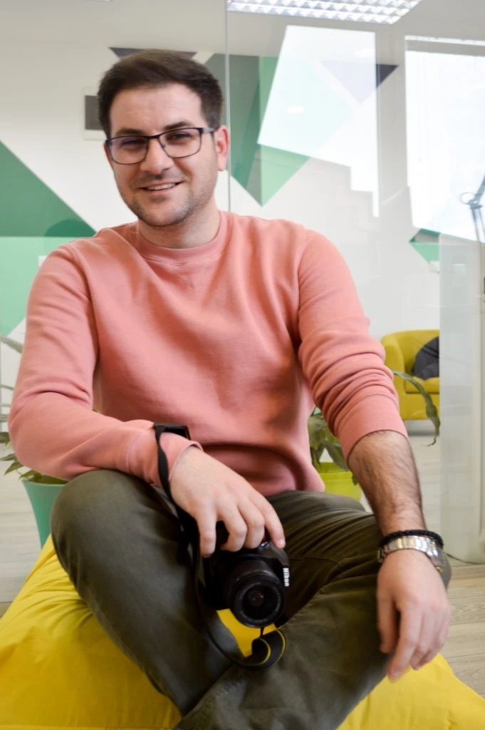 A person sitting on a bean bag in an office holding a camer and smiling